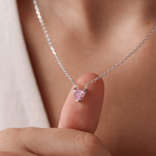 A close-up image of a sterling silver necklace with a white gold plating, featuring a dainty pink heart charm.