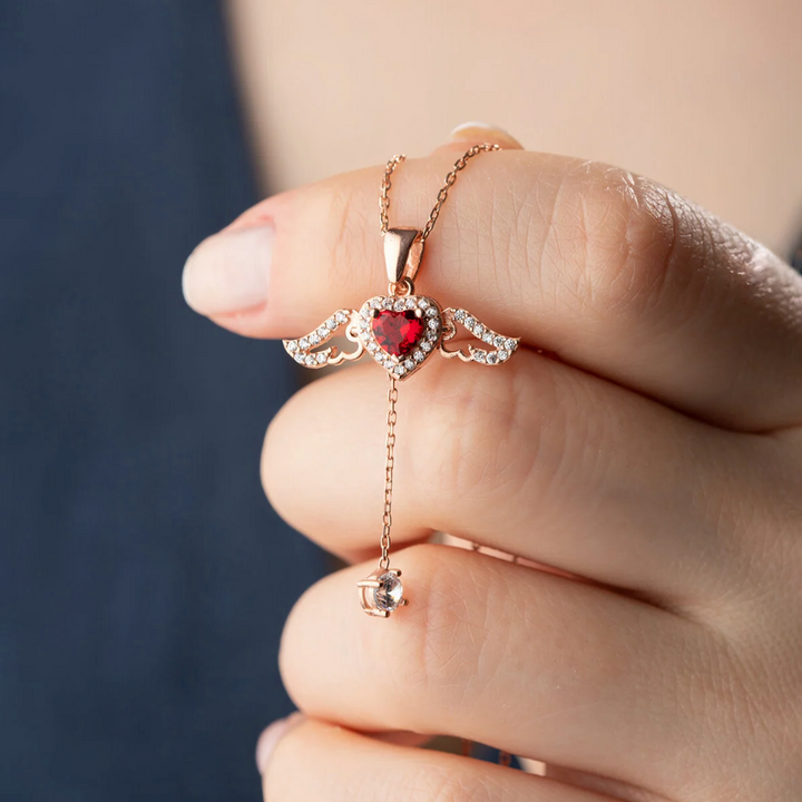 Rose gold necklace with a heart-shaped red gemstone held by a hand against a blurred background