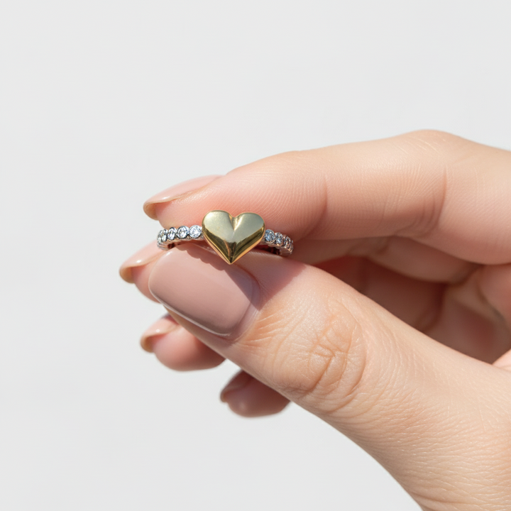 Hand wearing a gold heart-shaped ring with a clear gemstone on a light gray background