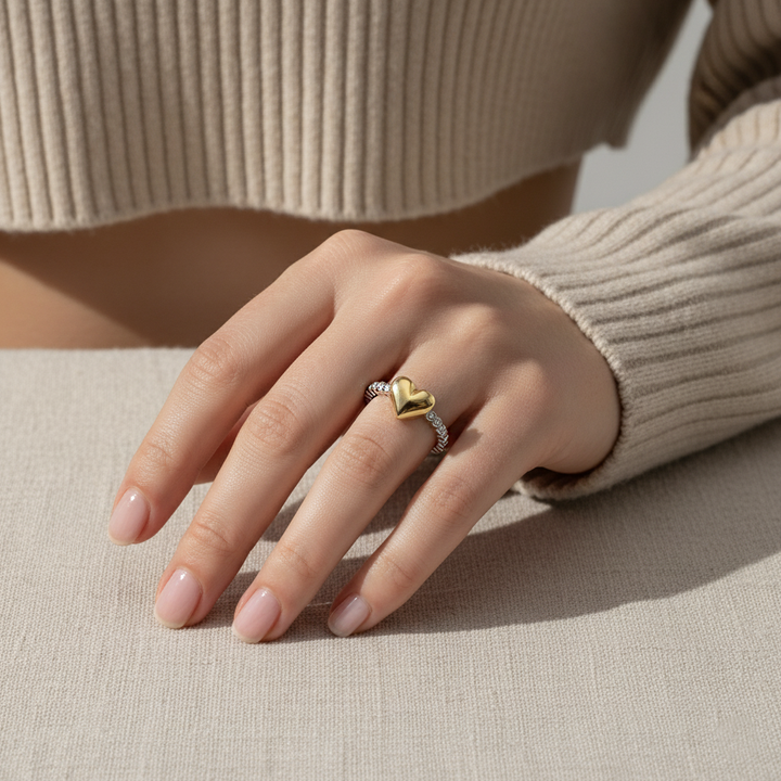Hand wearing a gold heart-shaped ring on a neutral background