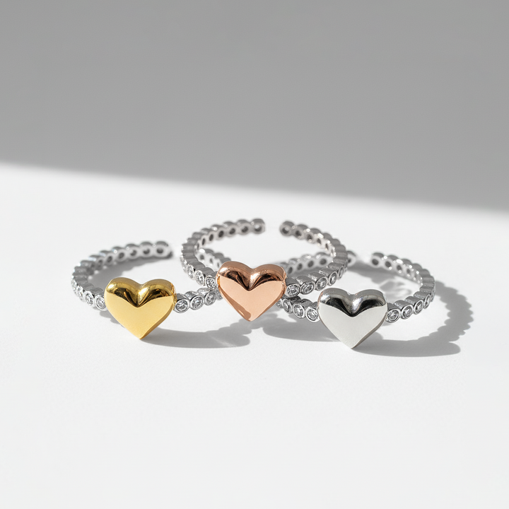 Three heart-shaped rings in gold, rose gold, and silver on a light gray background