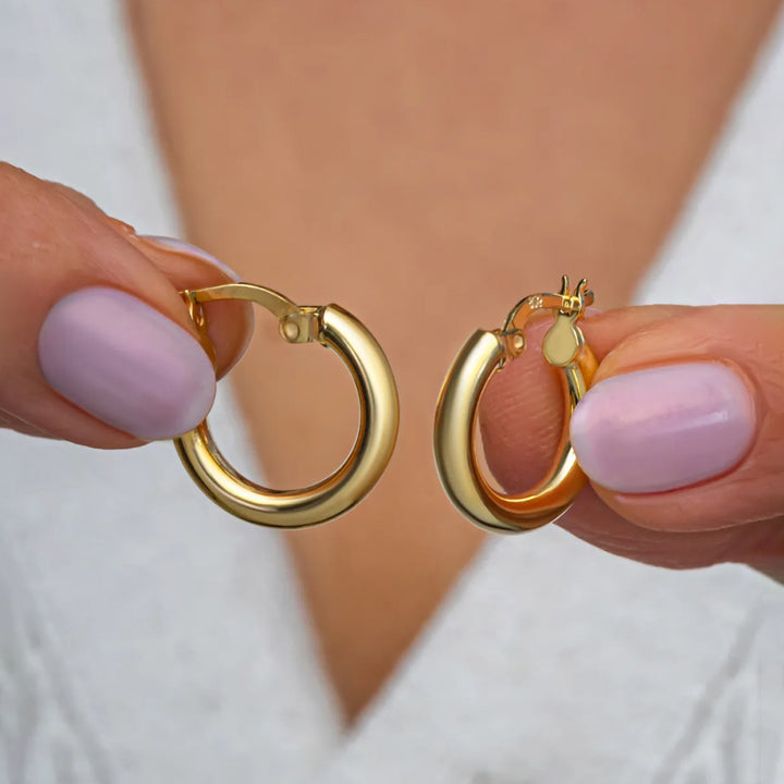 Gold hoop earrings held by a person with pink nail polish against a neutral background