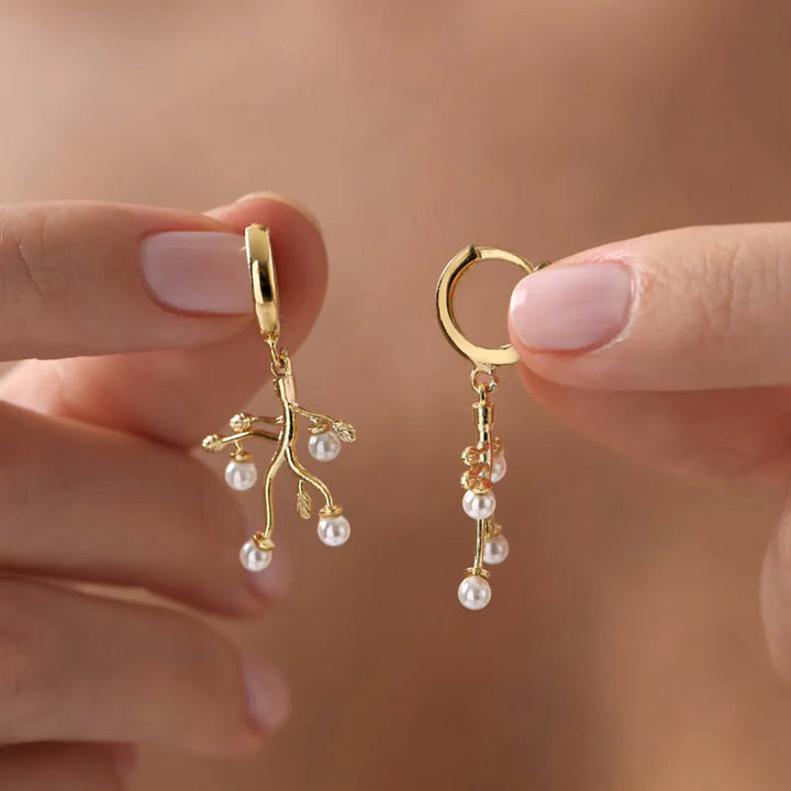 Gold earrings with pearl drops held between fingers against a neutral background