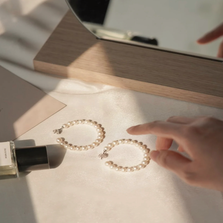 Pearl earrings on a table with a hand reaching towards it, next to a perfume bottle.